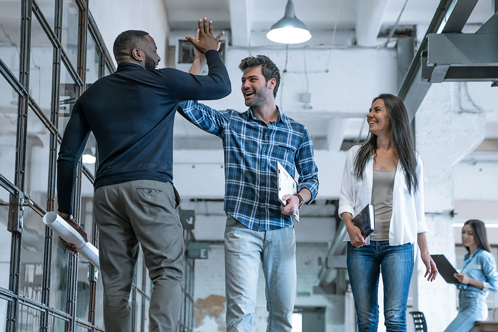 People in a common area of an office. Two people are giving a high-five while walking past each other.